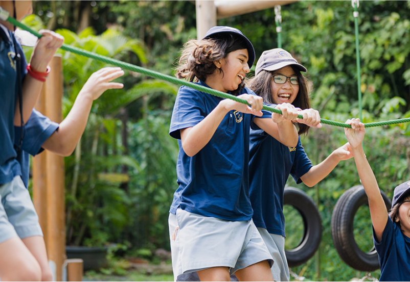 Two Invictus students in navy uniforms laugh together as they pull on a thick rope in an outdoor adventure playground, with lush greenery and hanging tyre swings behind them.