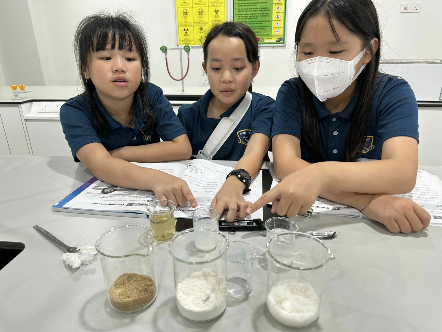 Three primary students at a lab bench pointing to beakers of powders and liquids, with open worksheets and safety posters behind them.