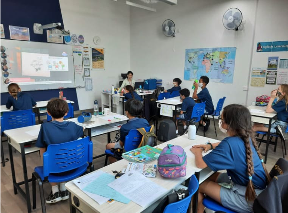 Primary students at Invictus Singapore sitting at socially-distanced desks in a classroom, wearing masks and watching a teacher present Chinese vocabulary on a large projection screen.