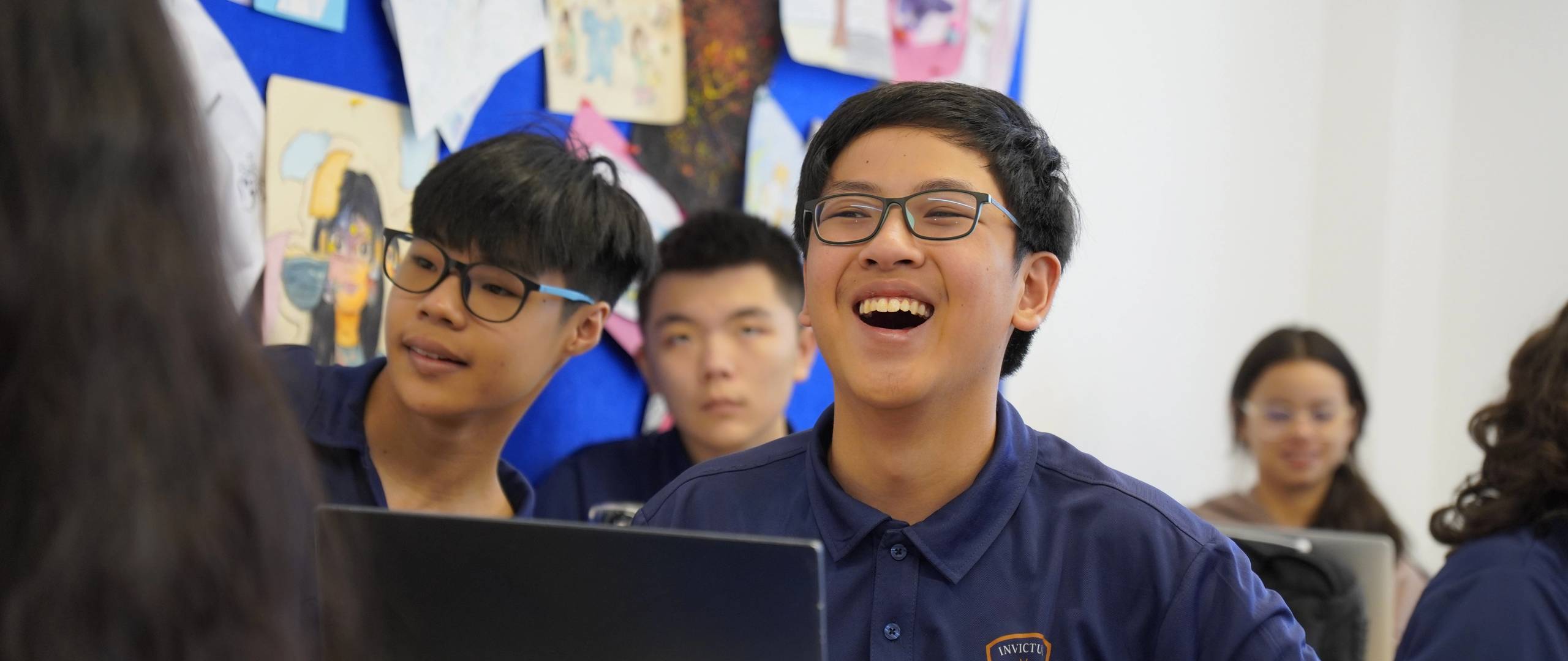 A group of Invictus International School Phnom Penh Year 4 students wearing masks and school uniforms enthusiastically perform in front of an art display and a teacher, with colourful values like "CARE" and "EXCELLENCE" painted on structural pillars in the background.