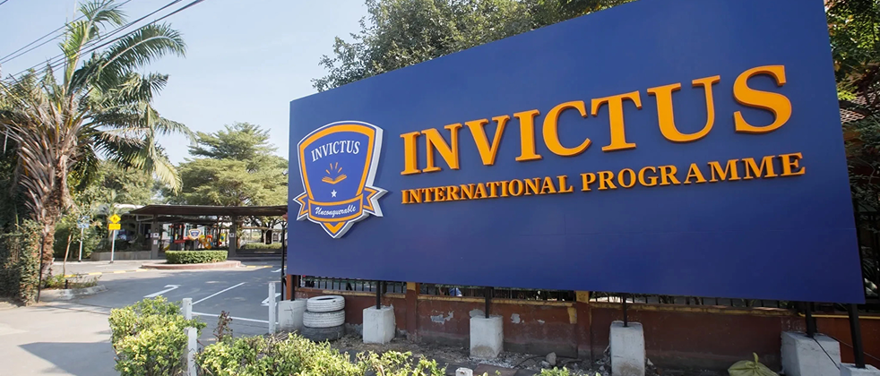 Large blue Invictus International Programme sign at the entrance of the Pathum Thani campus, surrounded by greenery and a clear driveway.