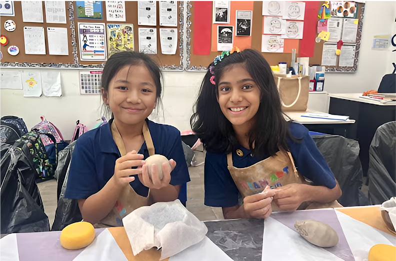 Two smiling students mould clay pieces in a creative pottery ECA, wearing protective aprons in a vibrant classroom setting.