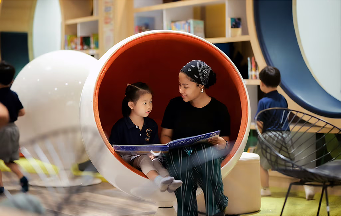 A teacher and young student sharing a book inside a modern, white-and-orange cocoon-style reading pod at Invictus Phnom Penh’s library, promoting a nurturing literacy environment.