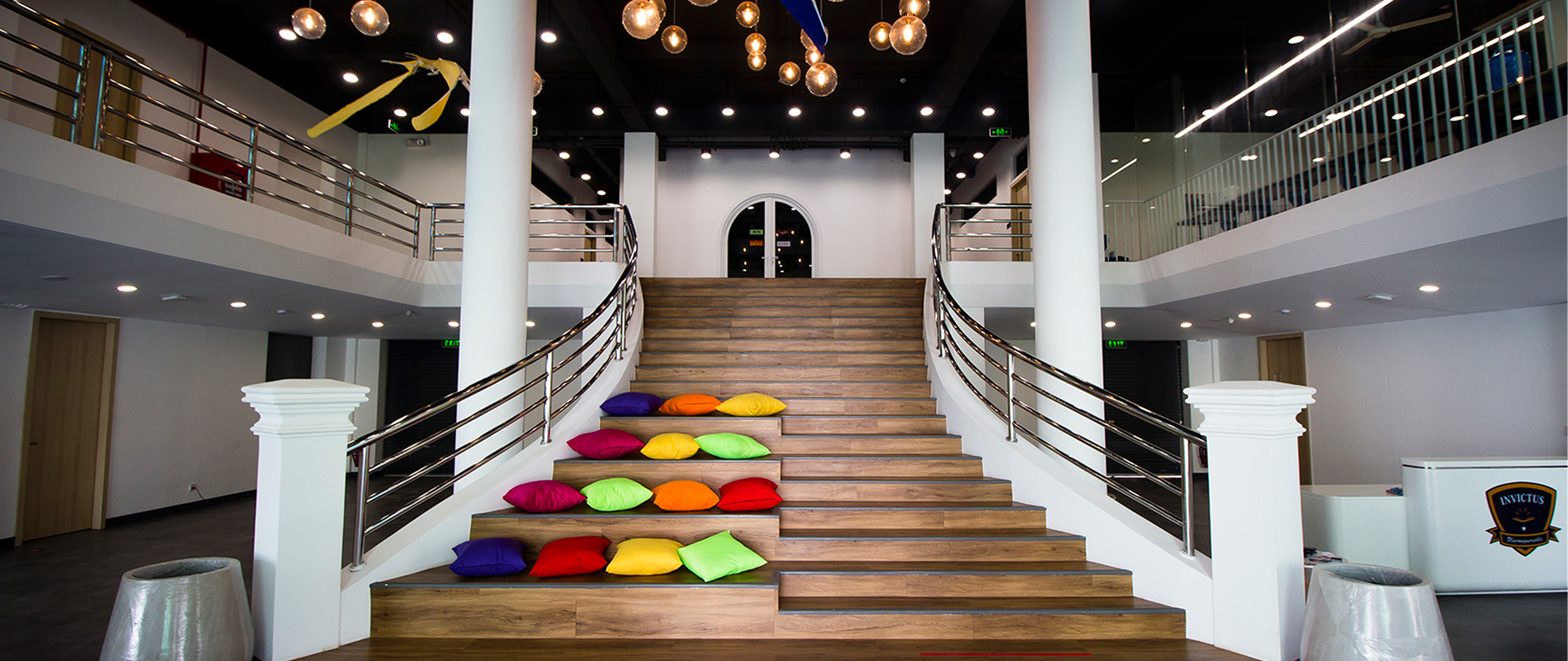 Grand staircase inside Invictus International School Phnom Penh, featuring wooden steps lined with colourful cushions for casual seating. The area is well-lit with modern hanging lights and surrounded by sleek white pillars, stainless steel railings, and a two-storey open-concept design with classrooms and glass-walled spaces above.
