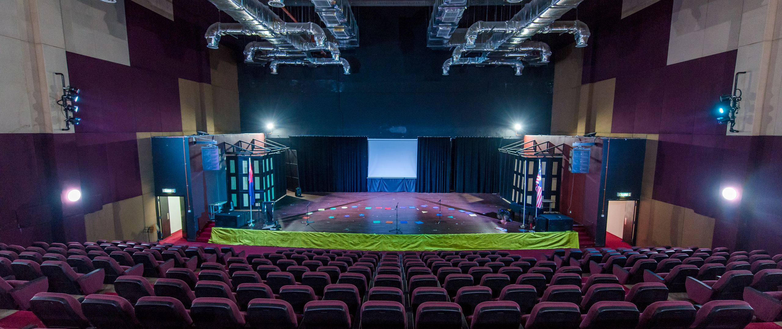 Wide-angle view of the Spring Hills Campus auditorium: tiered seating facing a stage under exposed ceiling ducts and theatrical lighting.