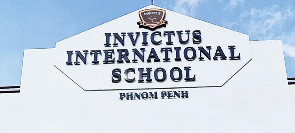 Invictus International School Phnom Penh’s white façade with the navy-blue and gold school crest and “INVICTUS INTERNATIONAL SCHOOL PHNOM PENH” lettering against a bright blue sky.