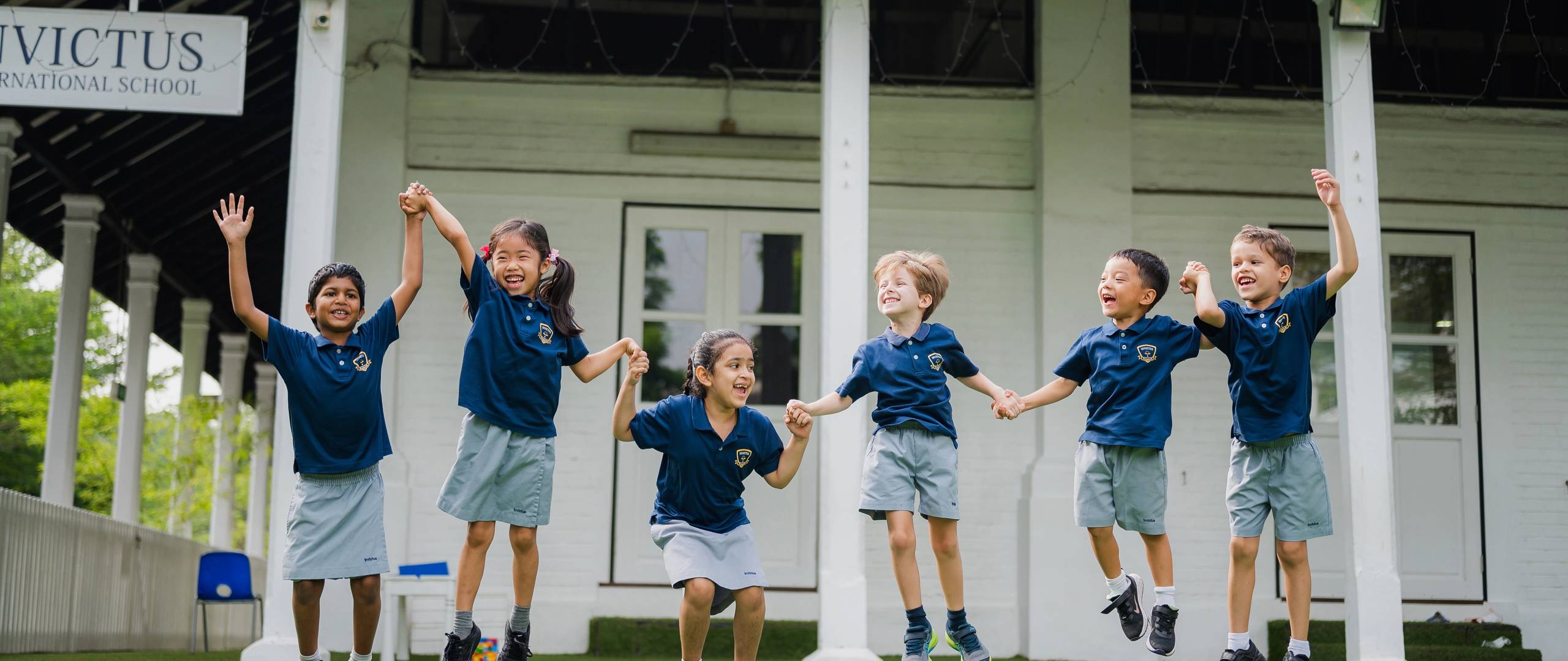 Group of young students in school uniforms holding hands and joyfully jumping together on a grassy field in front of an Invictus campus.