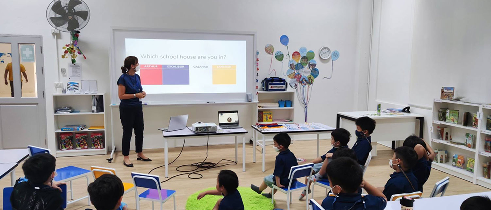 Primary students seated in a modern classroom at Invictus Pathum Thani, listening to a teacher present the school House system on a projector screen.