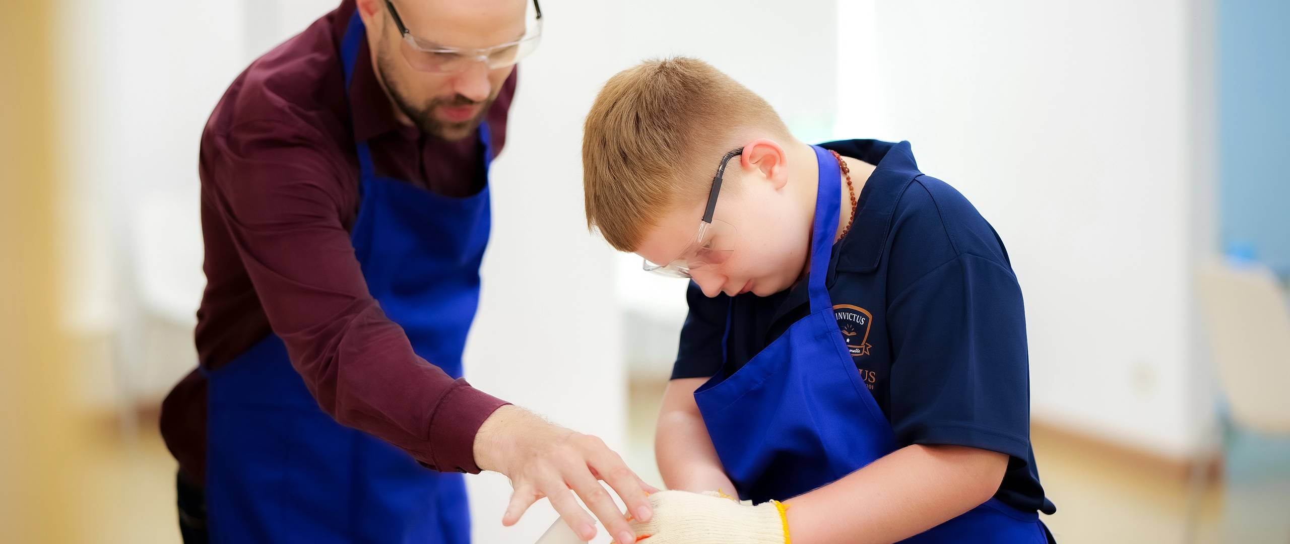 A group of Invictus International School Phnom Penh Year 4 students wearing masks and school uniforms enthusiastically perform in front of an art display and a teacher, with colourful values like "CARE" and "EXCELLENCE" painted on structural pillars in the background.