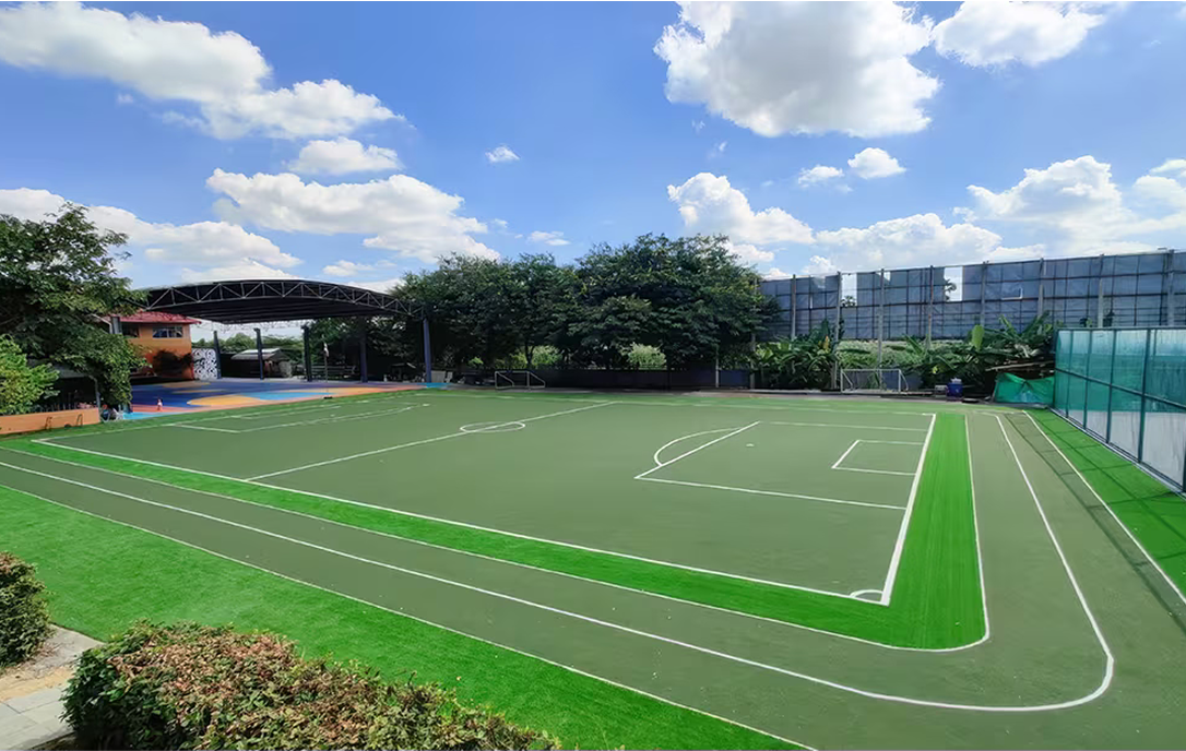 Spacious green artificial turf football field with surrounding running track under a sunny blue sky at Invictus Pathum Thani campus.