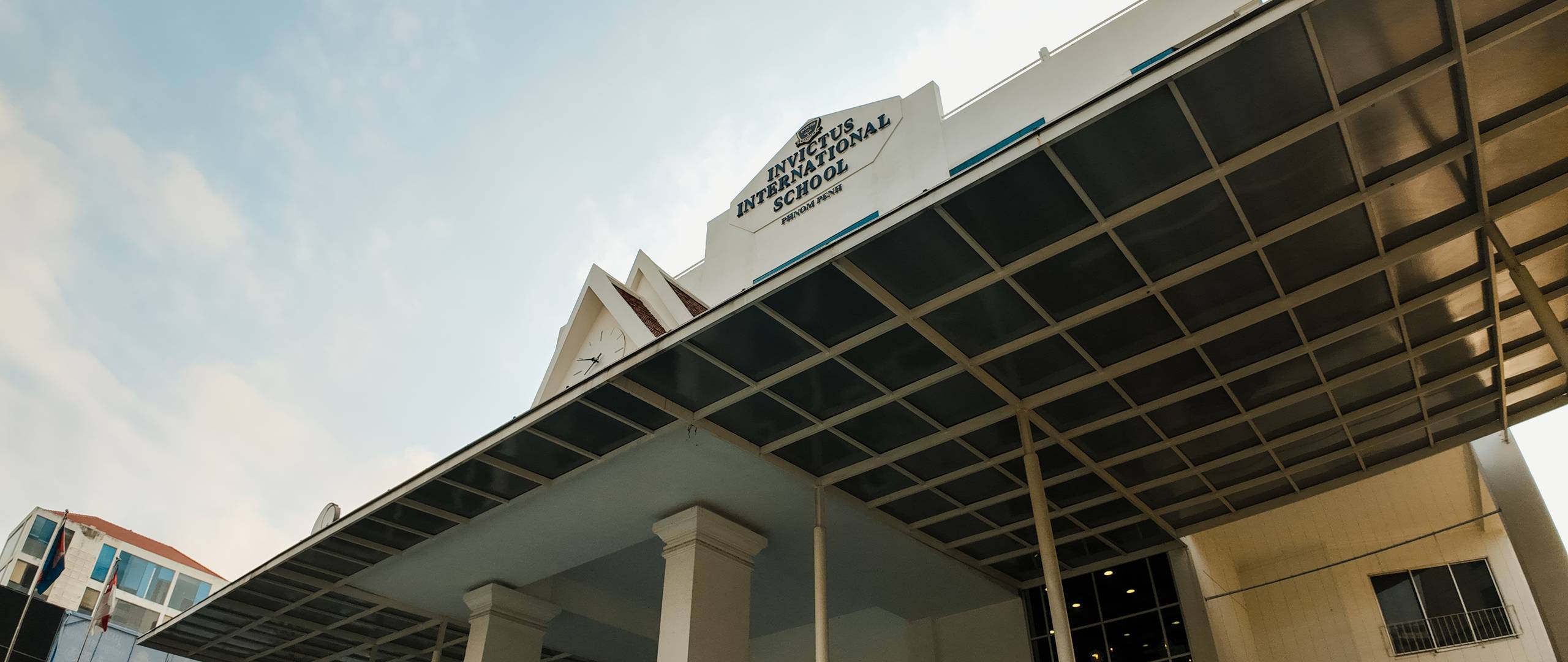 A group of Invictus International School Phnom Penh Year 4 students wearing masks and school uniforms enthusiastically perform in front of an art display and a teacher, with colourful values like "CARE" and "EXCELLENCE" painted on structural pillars in the background.