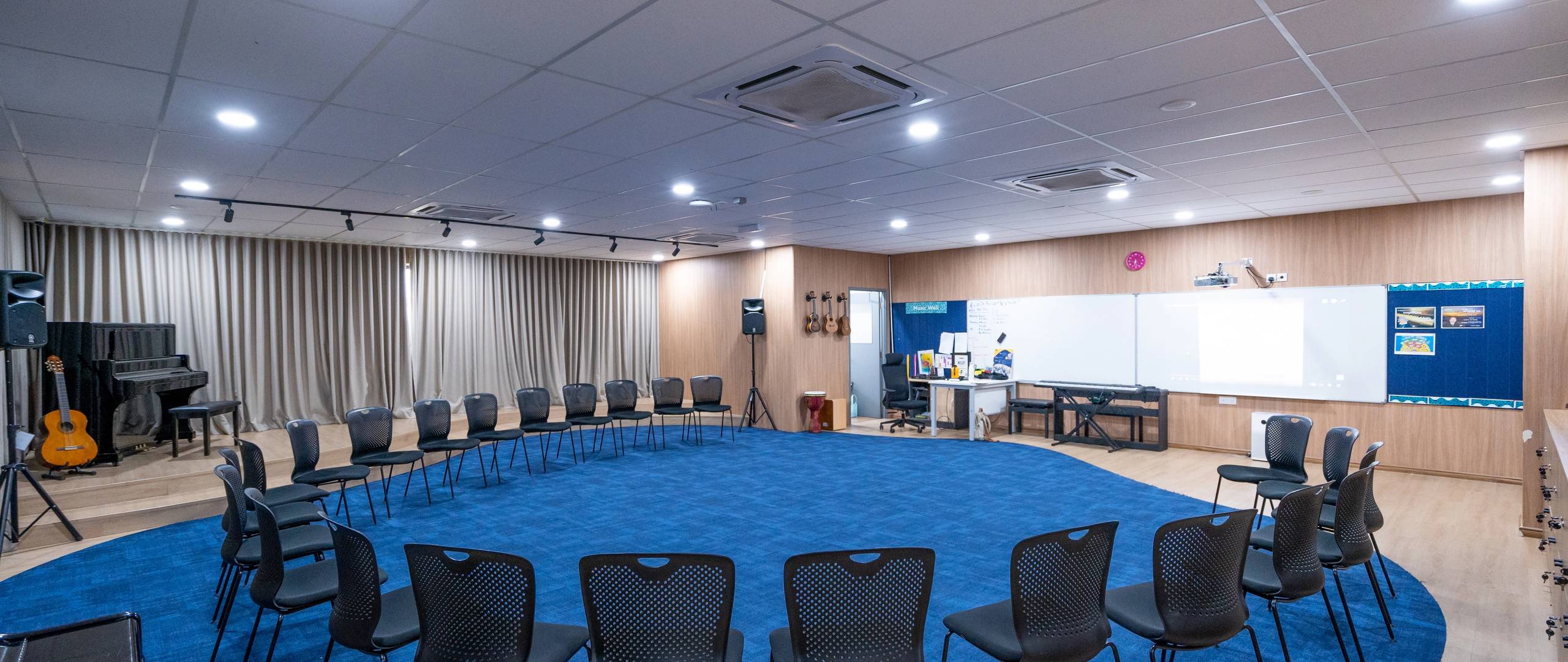 Music room with chairs in a circle, stage, instruments, and smartboard.