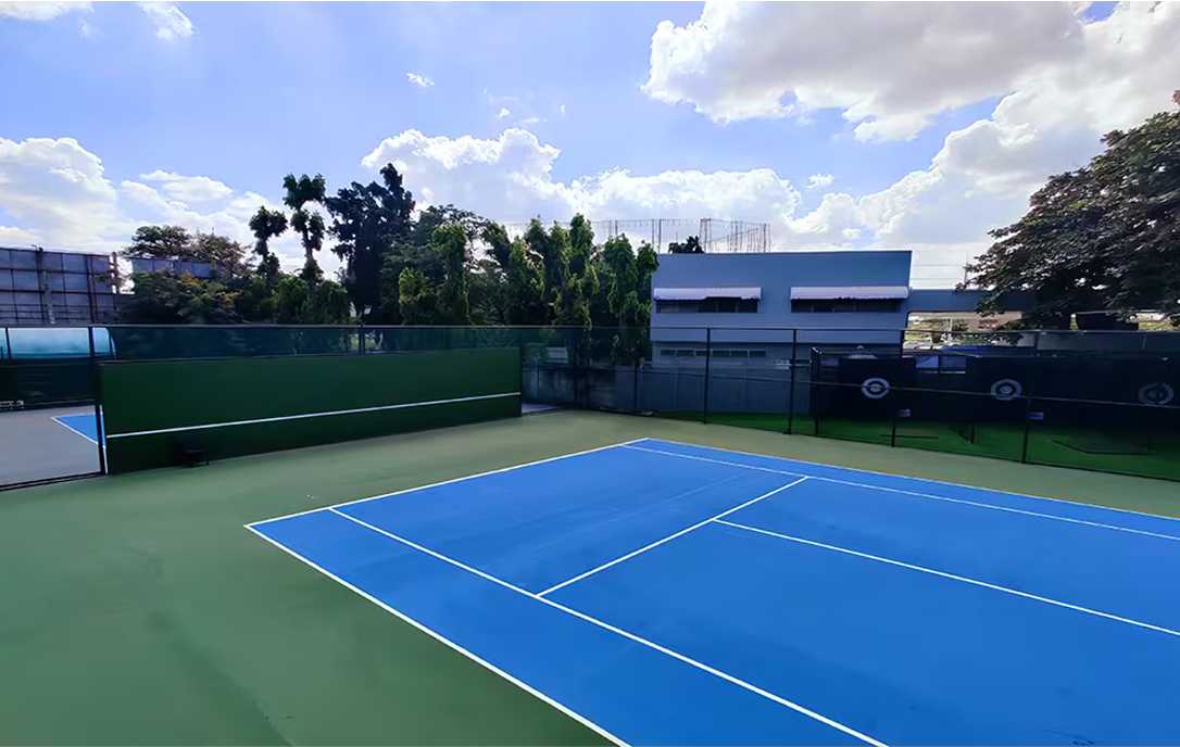 Bright blue tennis court with green borders, surrounded by trees and fencing, located at the Invictus Pathum Thani campus.