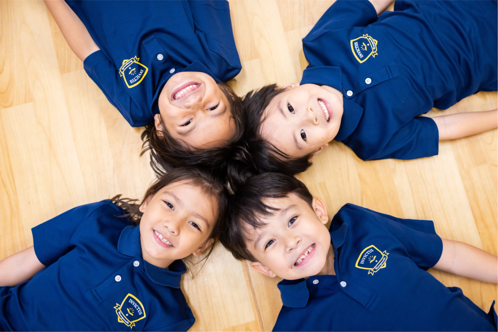 Top down view of four kids in blue uniforms laying on the wood floor, looking up and smiling.
