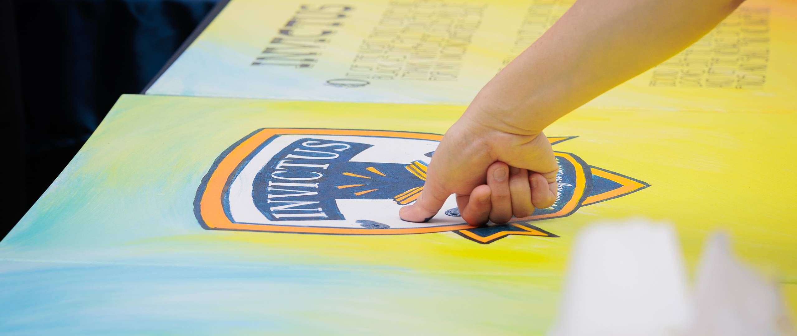A group of Invictus International School Phnom Penh Year 4 students wearing masks and school uniforms enthusiastically perform in front of an art display and a teacher, with colourful values like "CARE" and "EXCELLENCE" painted on structural pillars in the background.