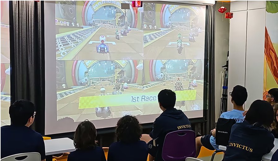 A group of secondary students wearing Invictus uniforms enjoy watching a Mario Kart tournament projected on a large screen during a gaming ECA.