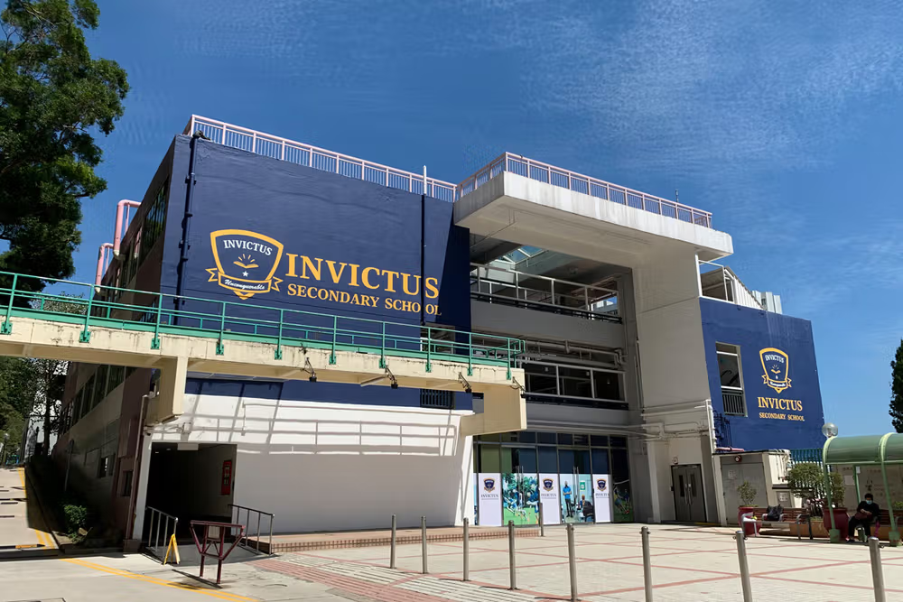 Exterior of Invictus International School’s Chai Wan Campus in Hong Kong, featuring a modern multi-storey secondary school building with navy-blue branding and a connecting walkway, under a bright blue sky.