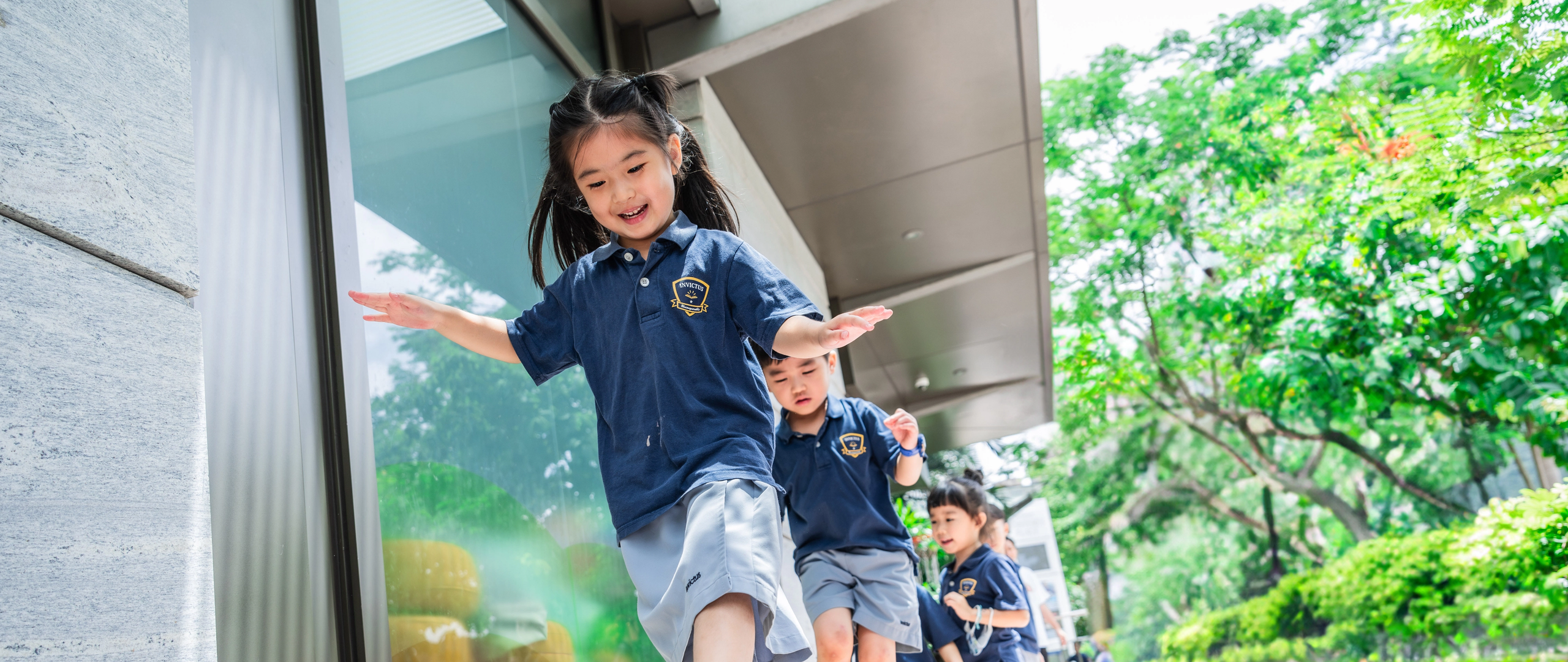 Three Invictus School Hong Kong students smiling and laughing outdoors under a sun umbrella, wearing school uniforms and hats.