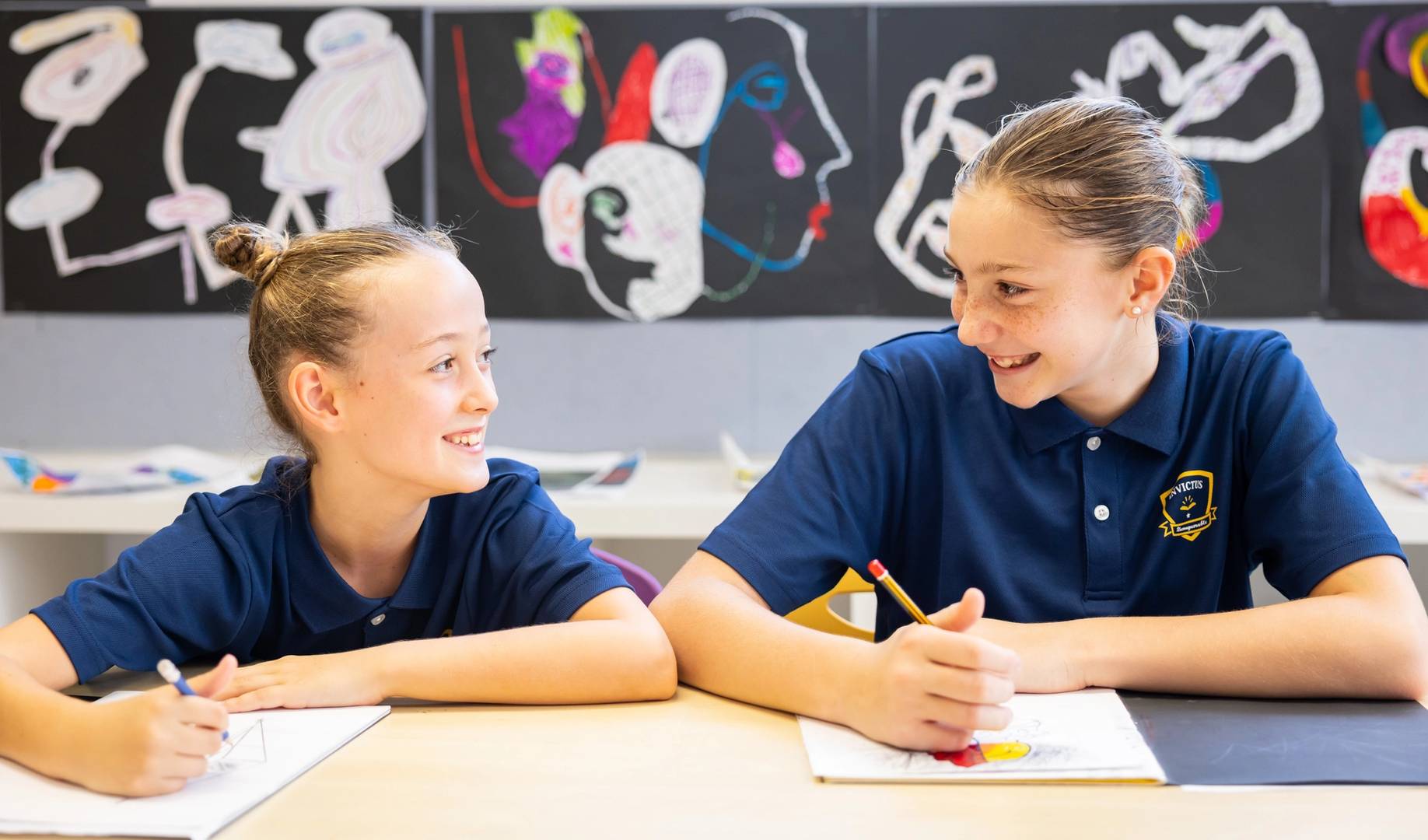 Two Invictus International School students smiling and engaging in an art activity together, seated at a table with sketchbooks and colourful drawings in a vibrant classroom adorned with abstract student artwork.