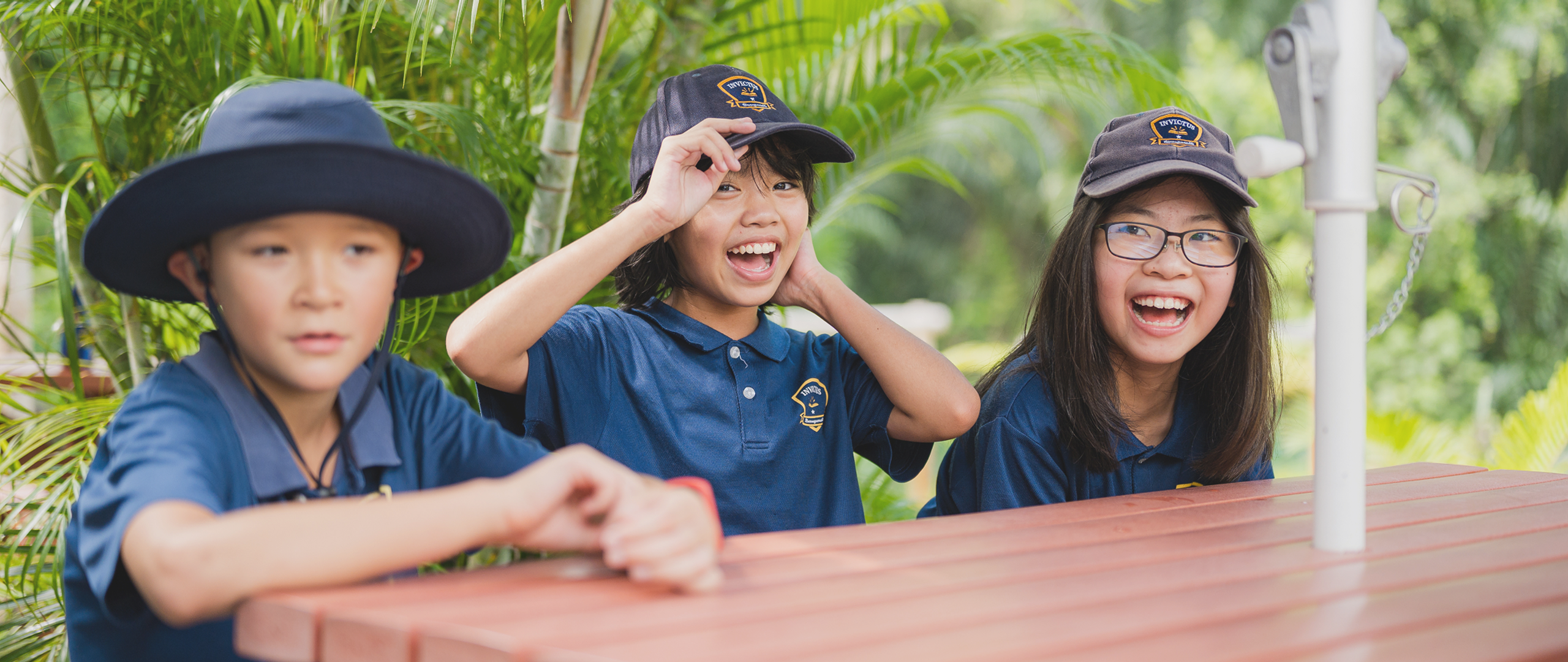 Three Invictus School Hong Kong students smiling and laughing outdoors under a sun umbrella, wearing school uniforms and hats.