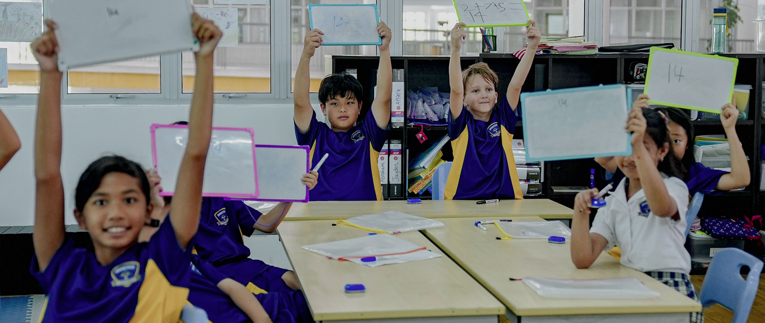 A circle of primary-aged students in purple and white Invictus uniforms seated around classroom tables, each proudly holding up a small whiteboard with their work on display