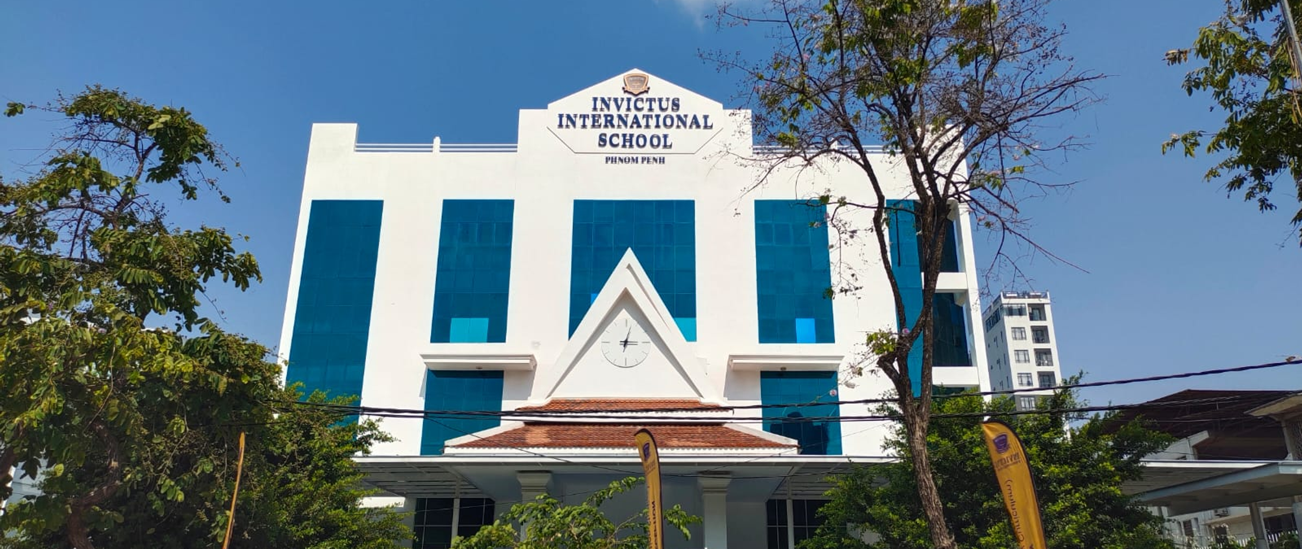 Front view of Invictus International School Phnom Penh campus with banners announcing open registration for Nursery to Secondary levels.