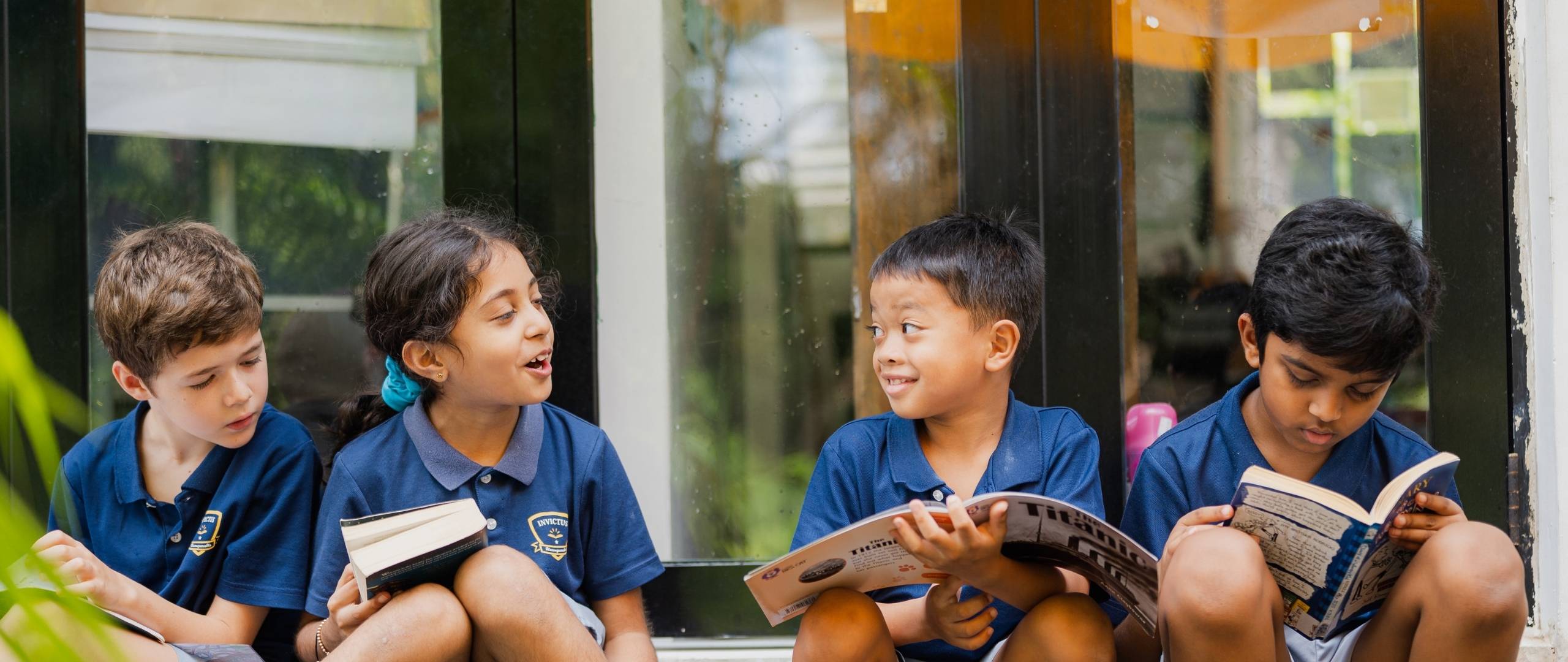 A group of Invictus Primary School students in uniform sitting outdoors, reading books and smiling together during a relaxed reading session.