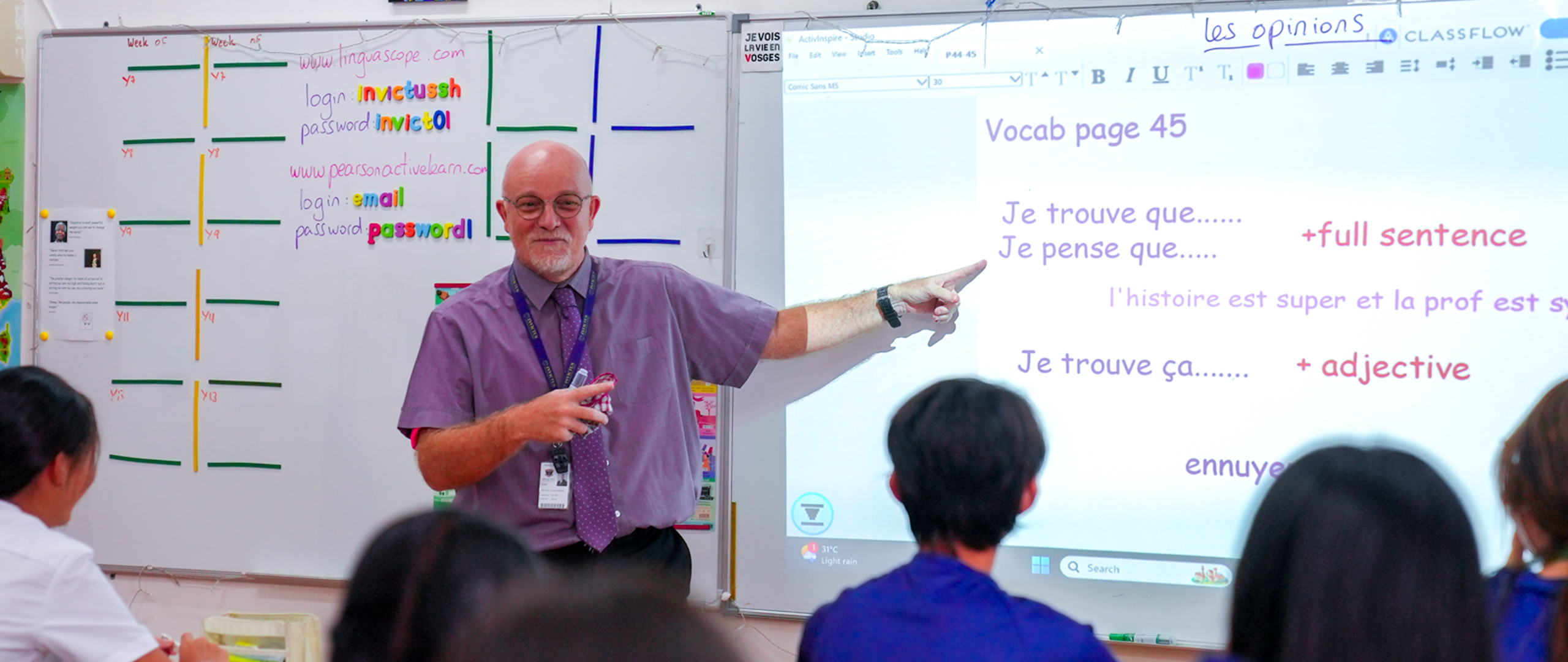 A middle-aged male teacher in a purple shirt and tie stands in front of a whiteboard and interactive display in a bright classroom, pointing to French vocabulary prompts (“Vocab page 45,” “Je trouve que…,” “Je pense que…”) while students sit facing him.