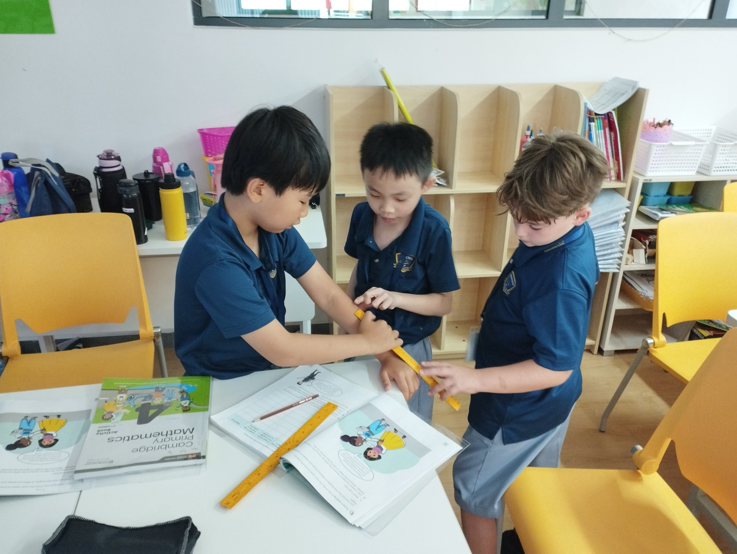 Three primary students in blue uniforms gather at a desk, using a yellow ruler to measure a classmate’s wrist over open math textbooks.