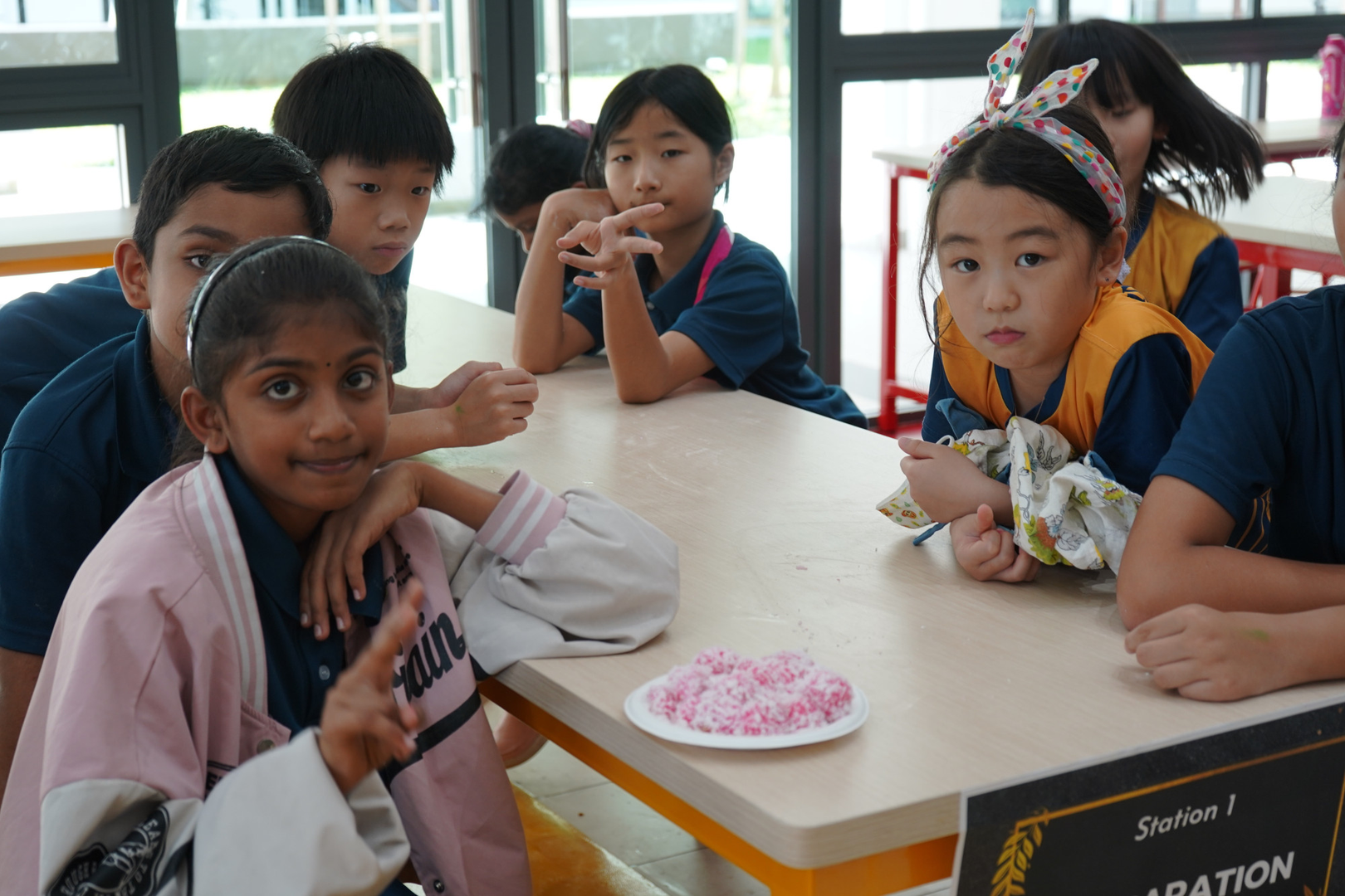 A group of primary students seated at a table labeled “Station 1,” gathered around a plate of pink coconut-coated treats, some looking at the camera.