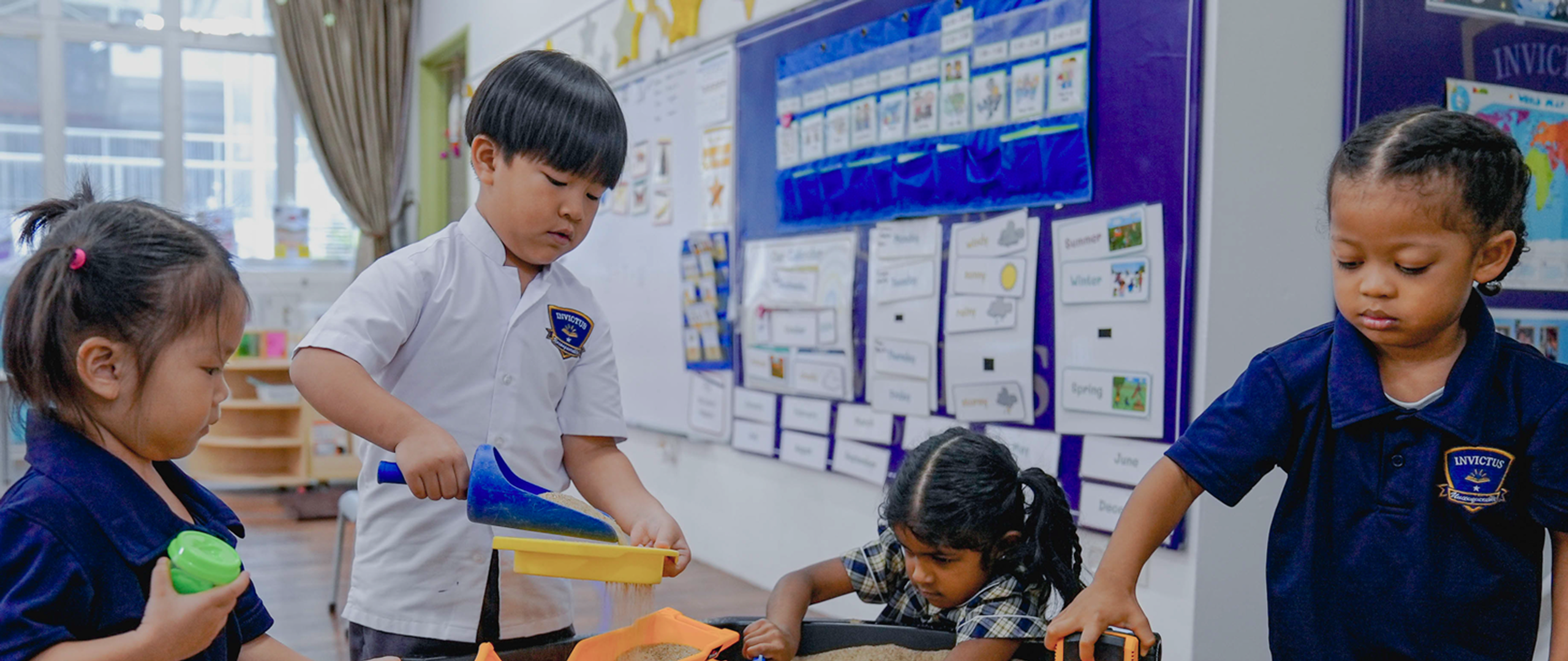 Children in Invictus Spring Hills Early Years classroom pouring and exploring sand in a sensory tray under teacher guidance.