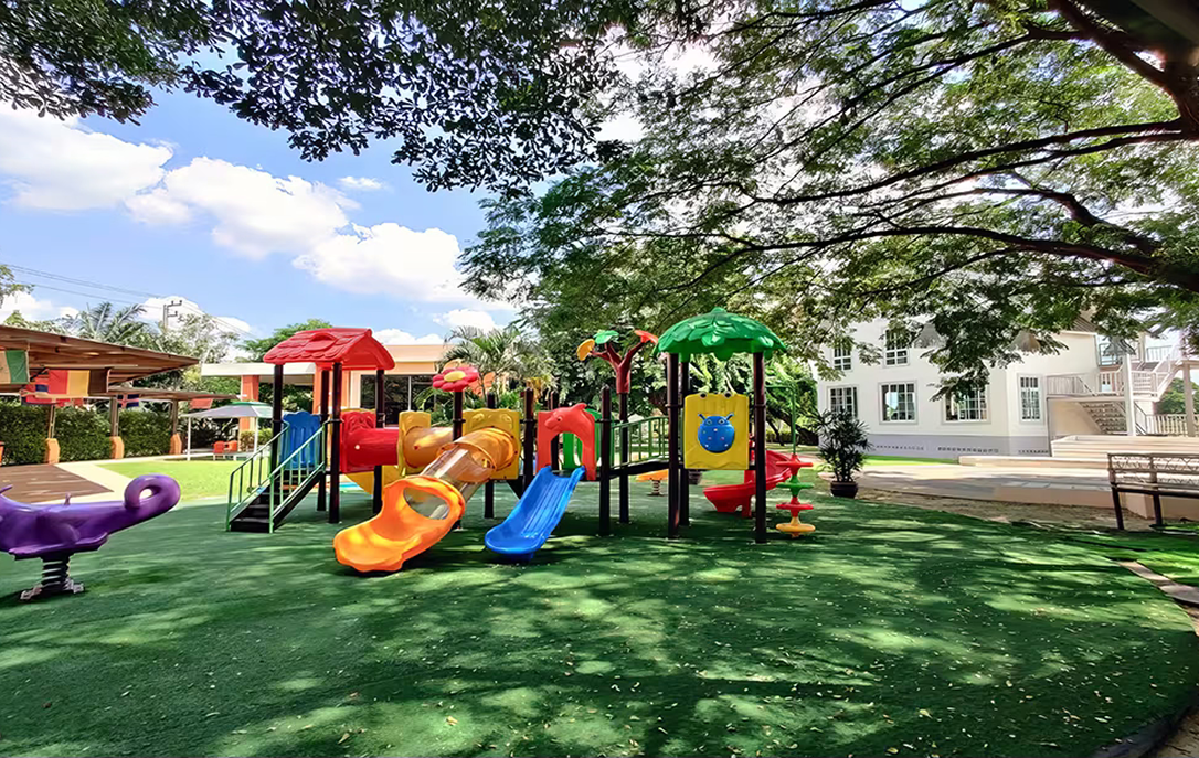 Colourful outdoor playground featuring slides, climbing structures, and activity panels set on a soft green turf beneath shady trees at Invictus Pathum Thani.