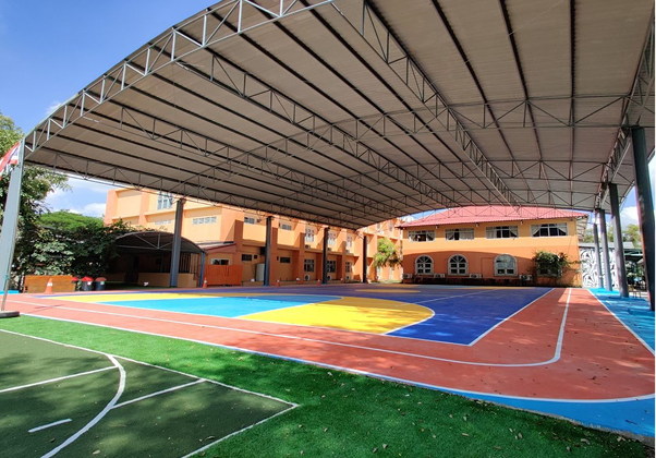 Covered multi-purpose sports court with vibrant blue, yellow, and red flooring, located beside the main school building at Invictus International School Pathum Thani, Thailand.