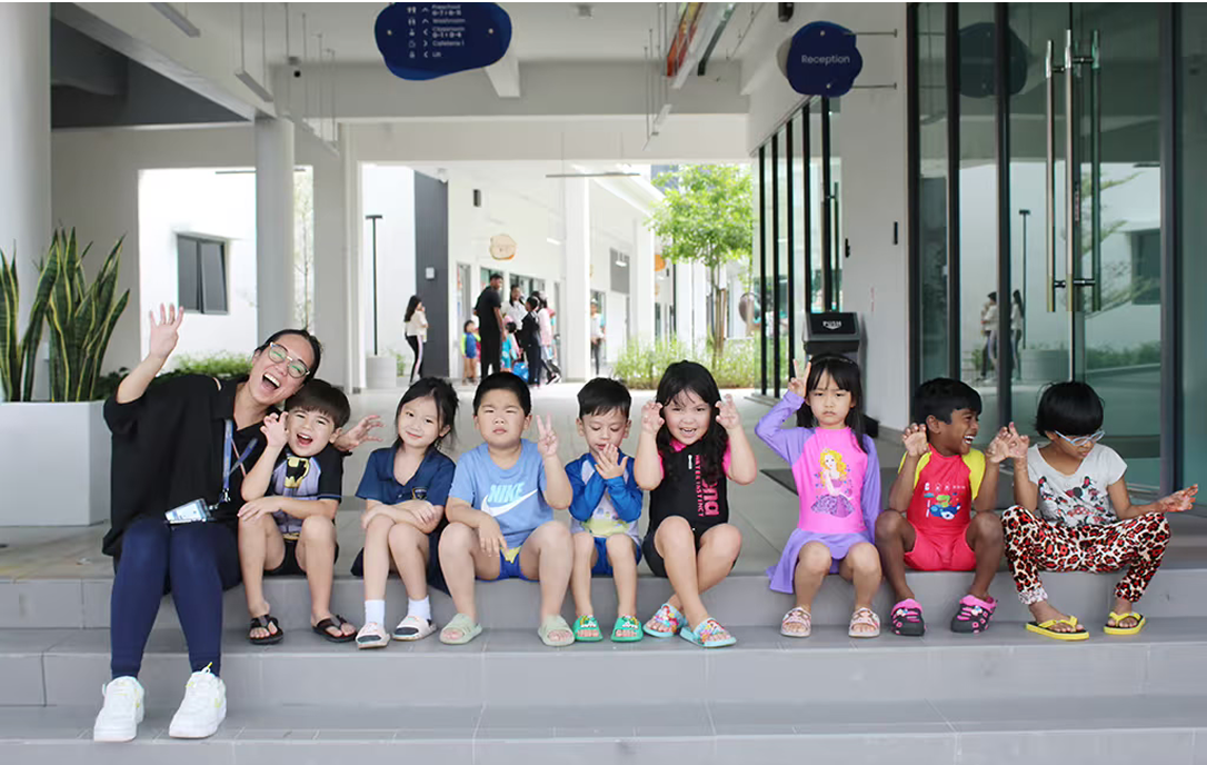 Group of young Invictus students and their teacher smiling and waving while seated outside the campus building, reflecting the joyful and inclusive school community.