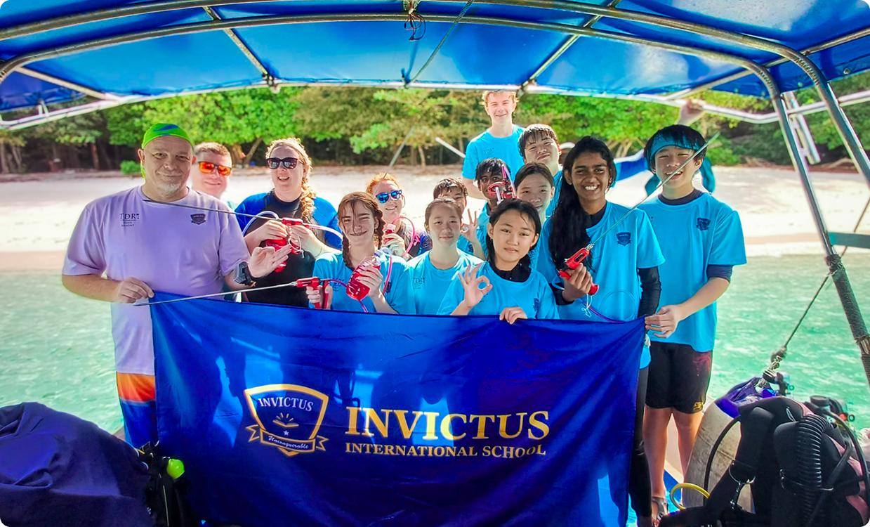 Invictus International School students and teachers on a boat, holding a school banner, preparing for a scuba diving session near a tropical beach.