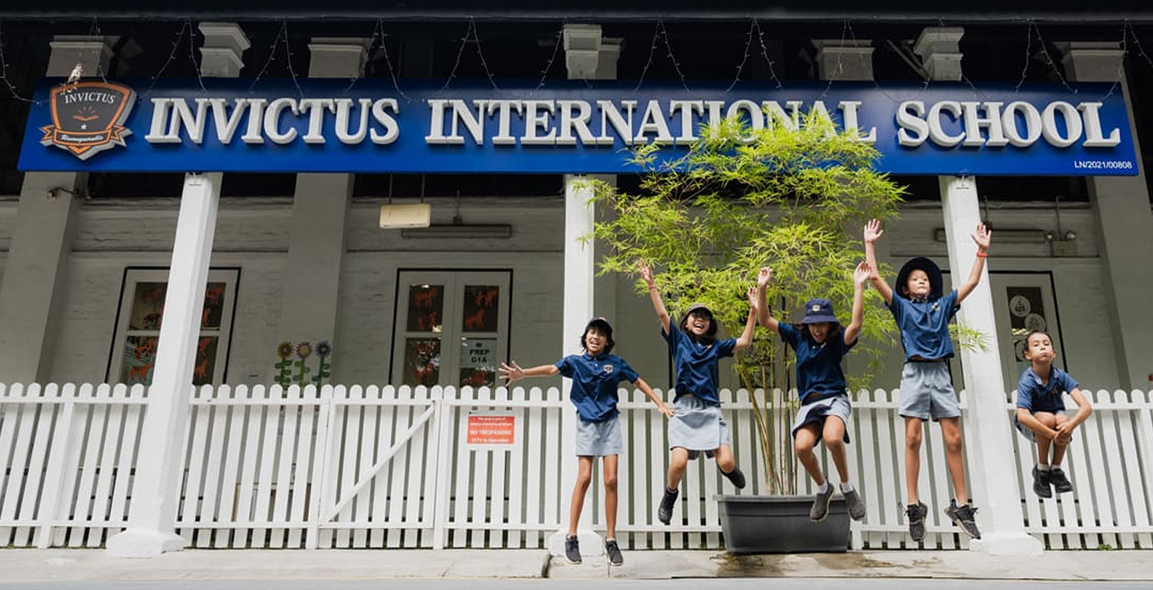 Group of young students in school uniforms holding hands and joyfully jumping together on a grassy field in front of a school building.