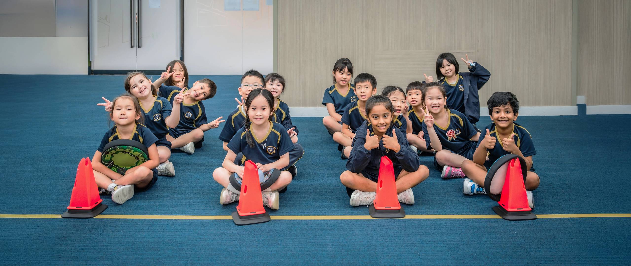 Group of young students in school uniforms holding hands and joyfully jumping together on a grassy field in front of a school building.