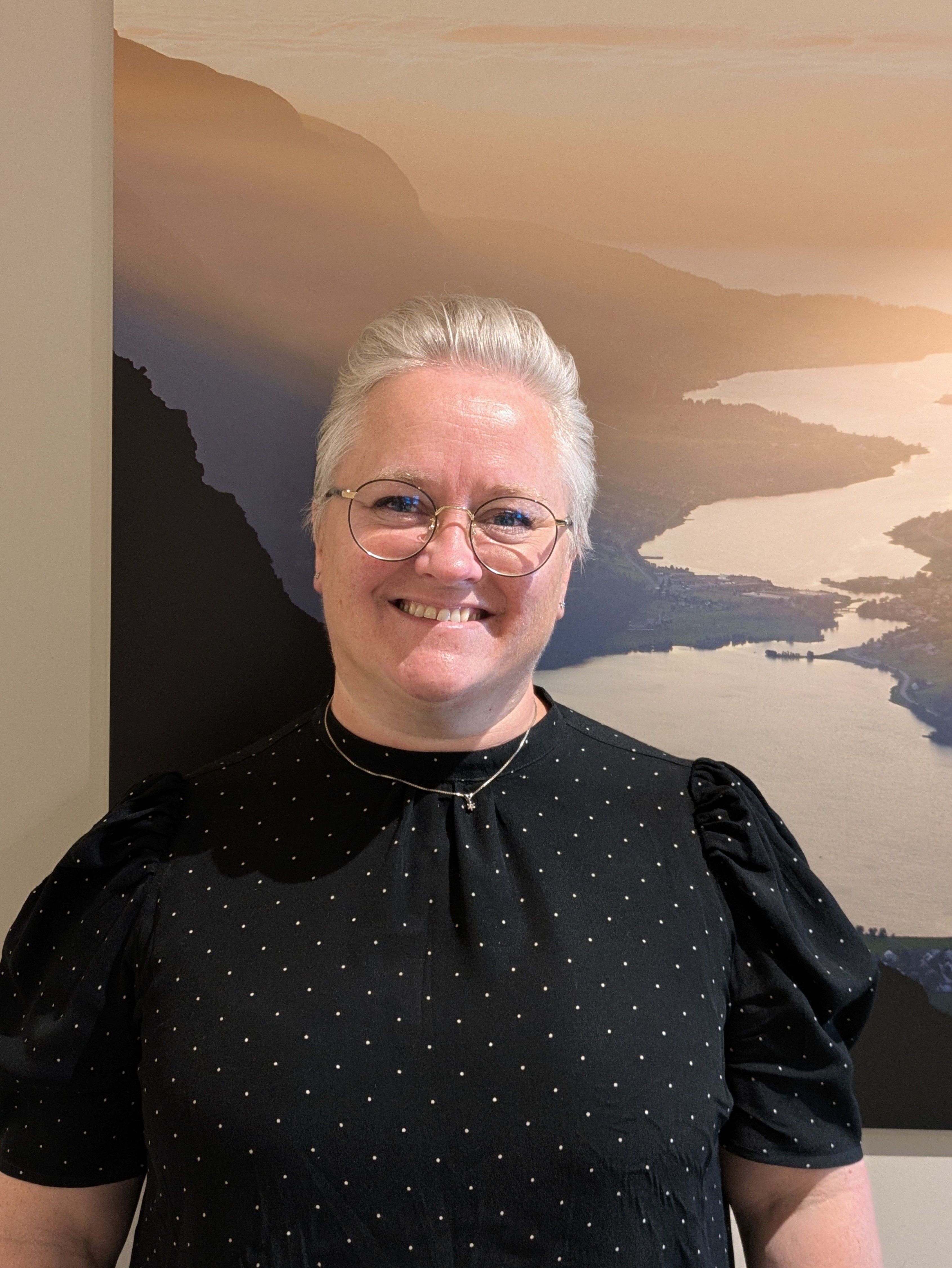 Bilde av smilende dame med kort lyst hår og briller, som står framfor eit utsiktsbildee av Sykkylvsfjorden