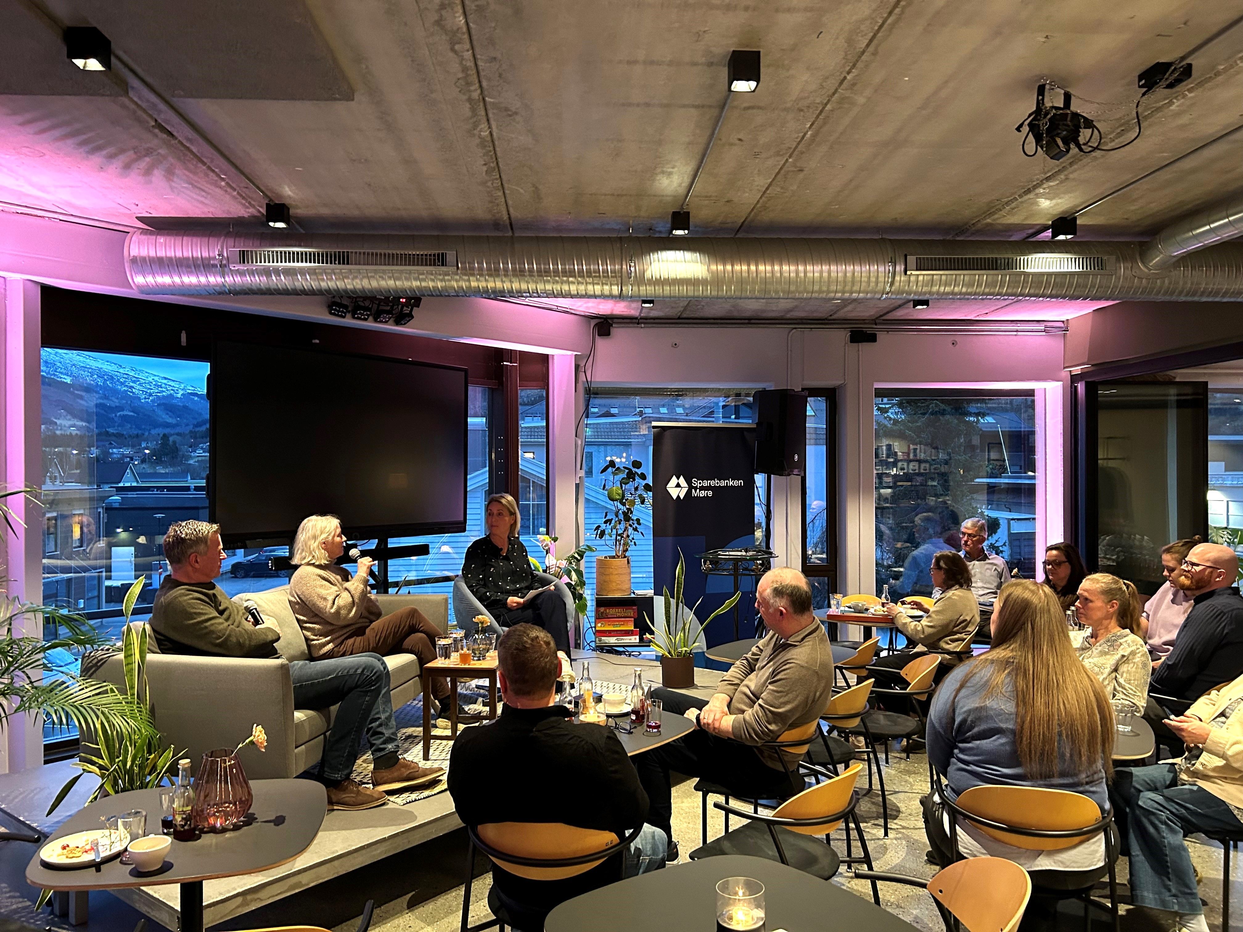 Bilde av 9 voksne mennesker i publikum, som lytter til 2 damer og en mann som sitter i samtale i en sofa på scenen