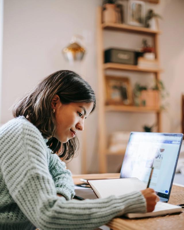 Estudante anotando no caderno, ao lado do notebook, em casa