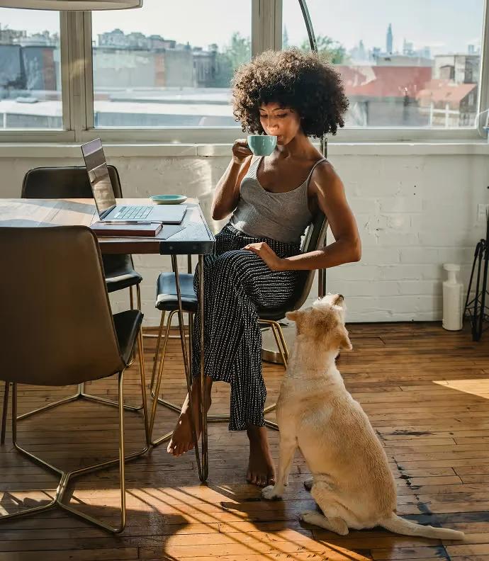 Mulher tomando café à mesa, diante do notebook, com um cachorro sentado ao lado
