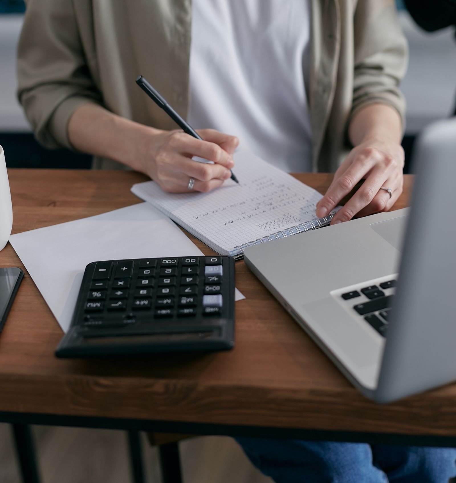 Mulher anotando contas num caderno, com calculadora e notebook na mesa