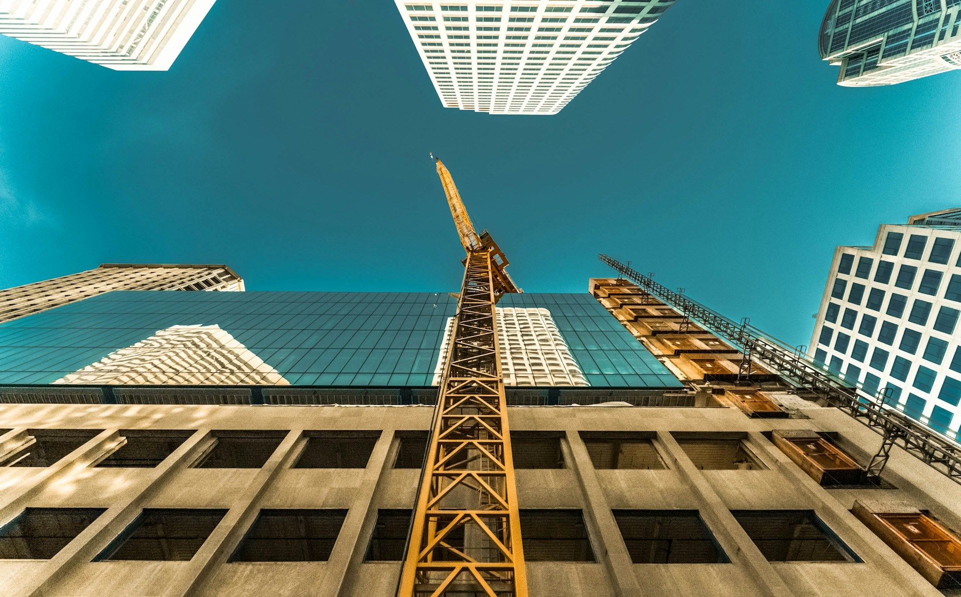 Yellow construction crane rising between skyscrapers against a teal sky