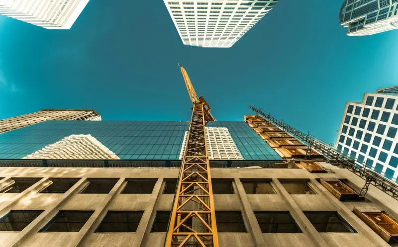 A low-angle photograph looking straight up at a construction site nestled between towering skyscrapers. In the center, a bright yellow construction crane rises vertically toward a clear, teal-colored sky.