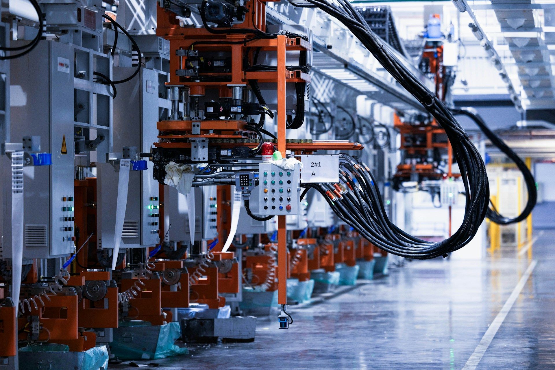 A high-angle, wide-perspective shot of an automated industrial assembly line inside a clean, modern factory. The scene is dominated by heavy machinery with bright orange structural frames and grey control panels.