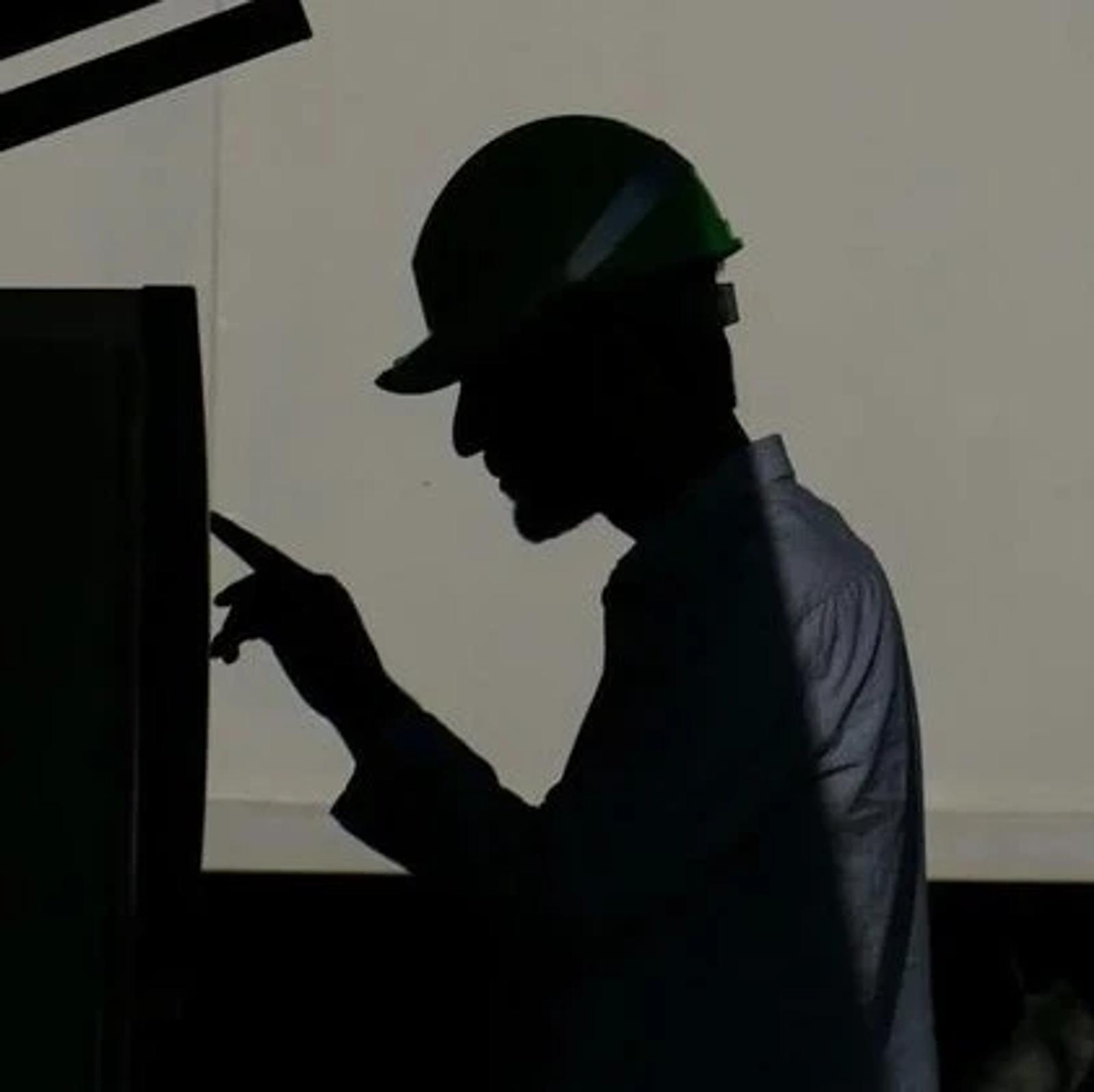 A worker in a hard hat silhouetted against a bright wall while operating a control panel