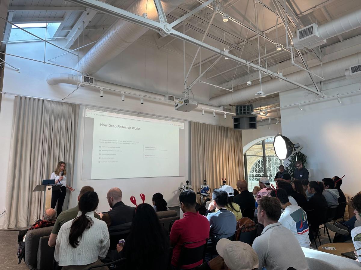 An audience sits watching a woman discuss a powerpoint presentation.