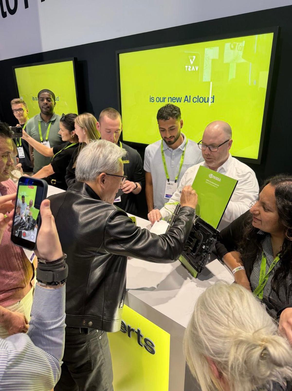 A man in a leather jacket, Jensen Huang, signs a computer screen while people watch