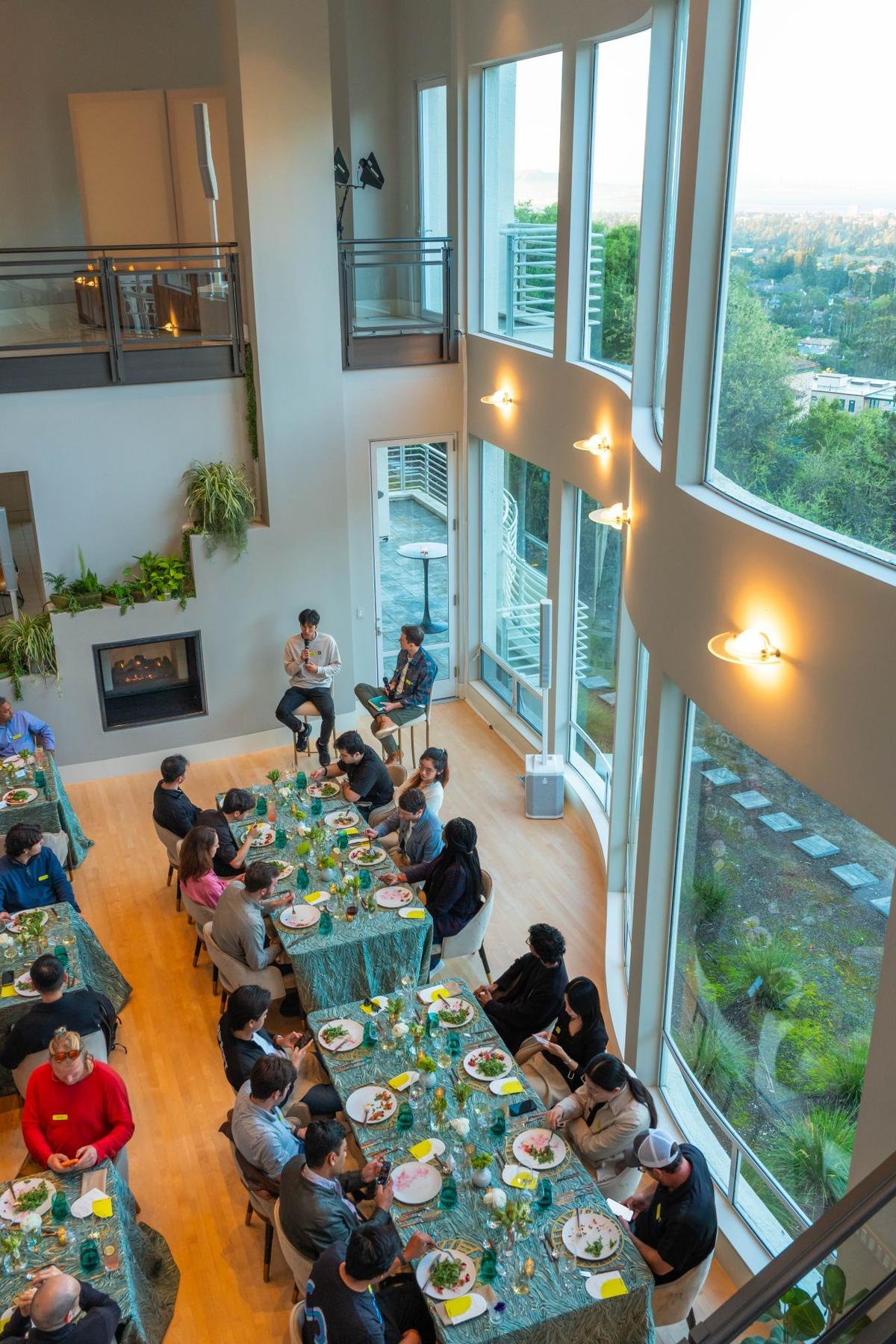 Lots of people sit at long tables with plates in front of them. Two people are on stools in the corner speaking to the audience.