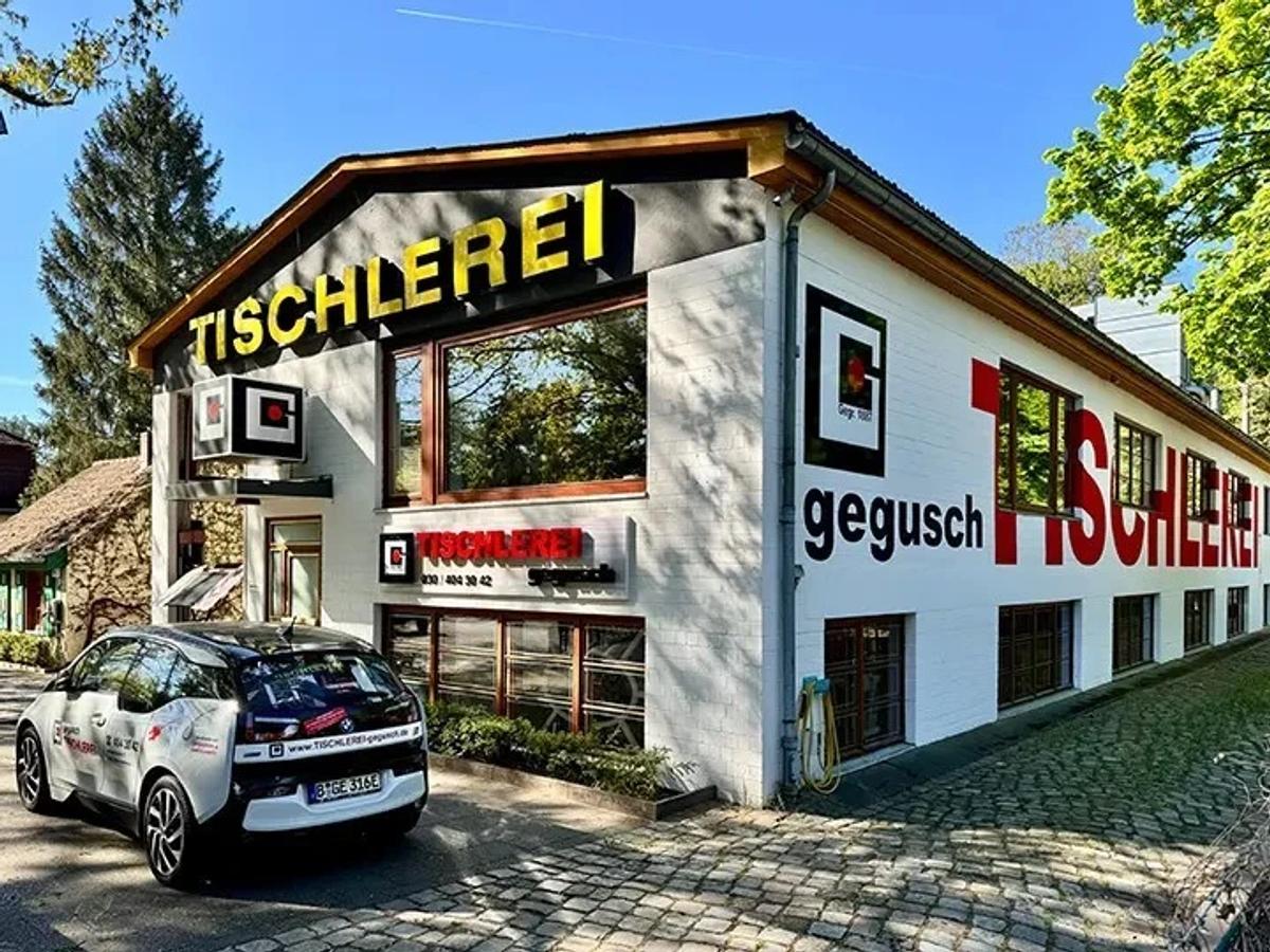 Das Werkstattgebäude der TISCHLEREI gegusch in der Hohefeldstraße: weiß gestrichene Klinkerfassade mit großem roten Schriftzug „TISCHLEREI", davor ein Elektroauto mit Firmenbeschriftung